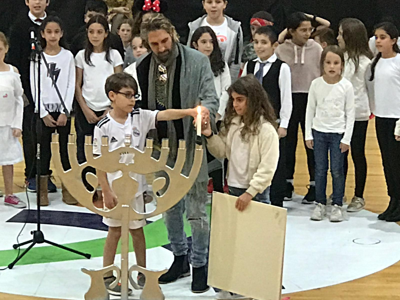 מוטי דנוס בטקס הדלקת נרות חנוכה בבית ספר, עם שני ילדים מדליקים את הנר